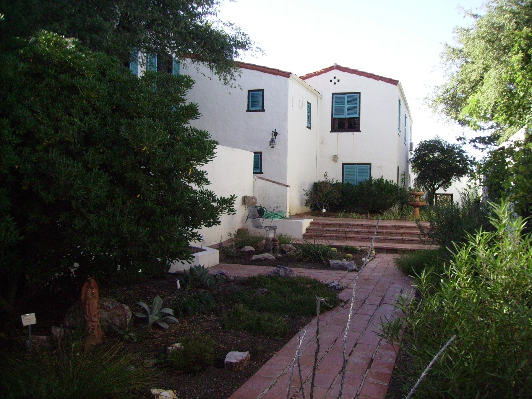 White Kannally Ranch House with green shutters, stepped brick paths and trees around its courtyard.