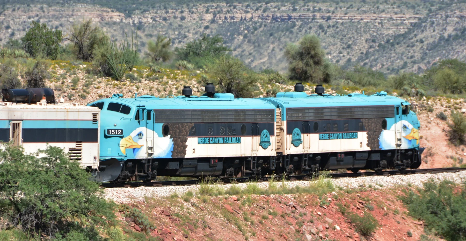 Two turquoise Verde Canyon Railroad locomotives beside desert vegetation and a rocky slope.