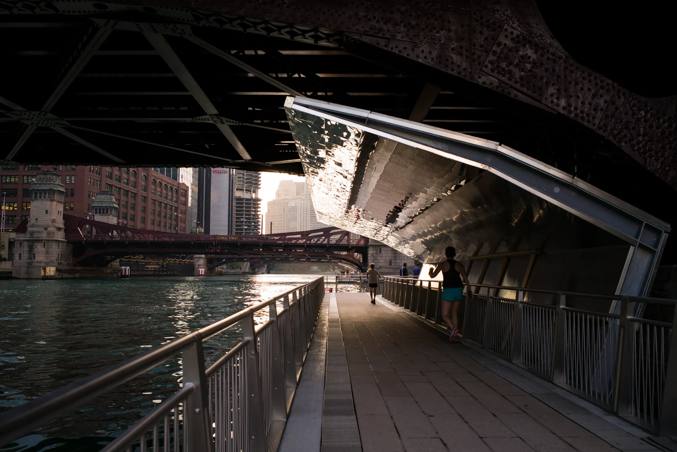 A riverside pedestrian path passes beneath a steel bridge toward sunlit buildings and another bridge.