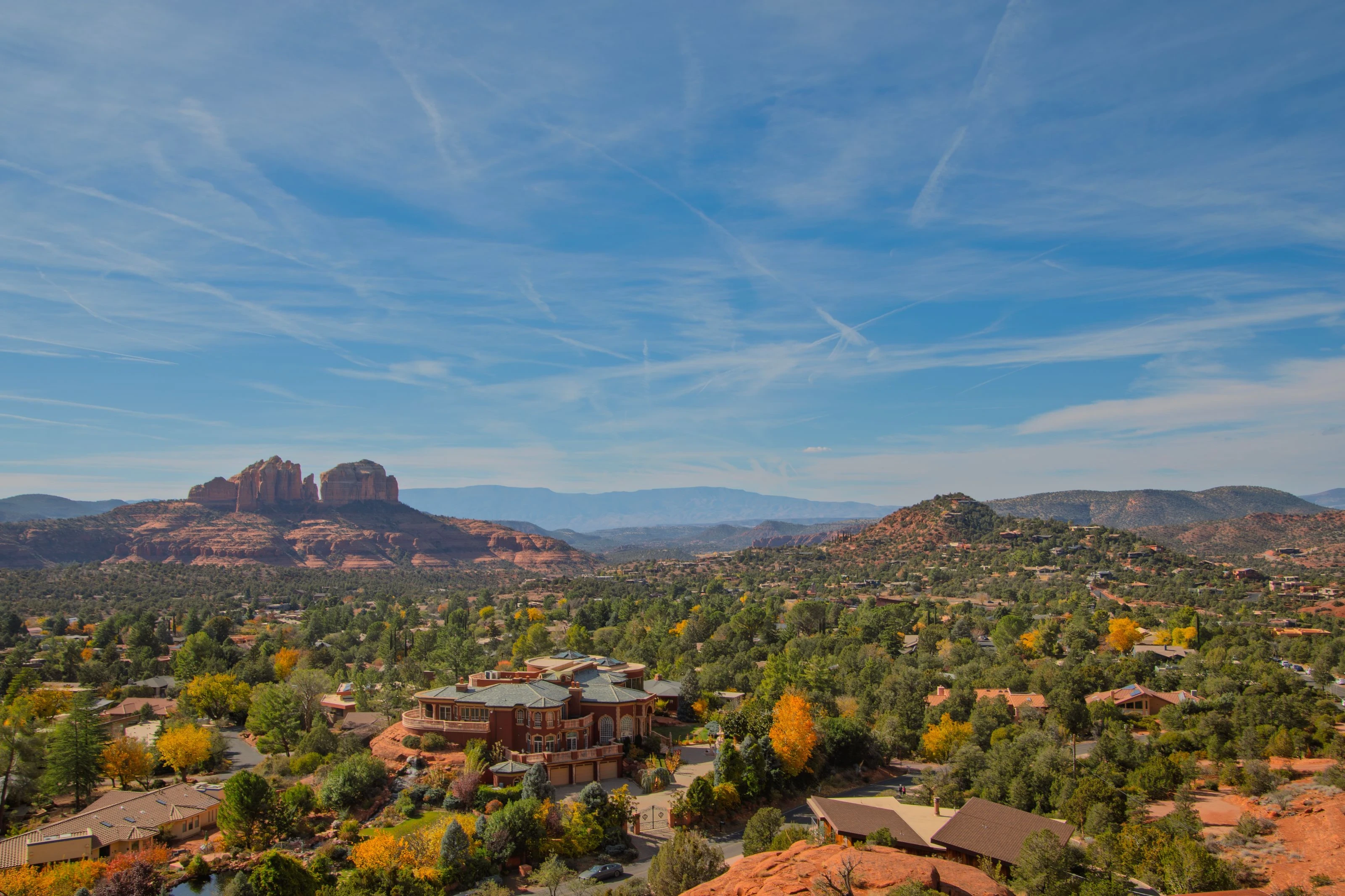 Sedona hillside homes among autumn trees with Cathedral Rock and distant red-rock mesas beneath a blue sky